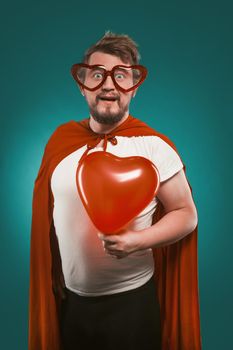 Superman In Love Holds Big Red Heart. Positive Man In Superhero Costume And Heart-Shaped Glasses Poses On Biscay Green Background. Valentine's day concept.