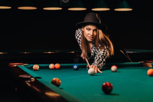 A girl in a hat in a billiard club with a cue in her hands hits a ball.Playing pool.