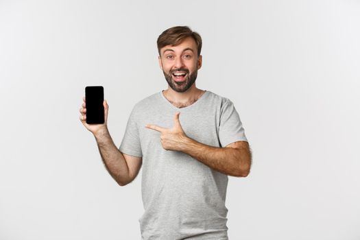 Portrait of excited man with beard, pointing finger at mobile phone screen and smiling, showing application, standing over white background.