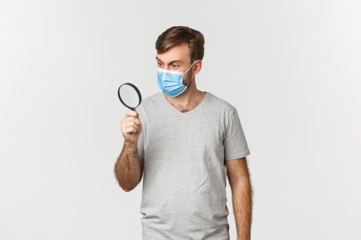 Concept of pandemic, covid-19 and social-distancing. Image of surprised man in medical mask, searching for something and looking through magnifying glass, standing over white background.