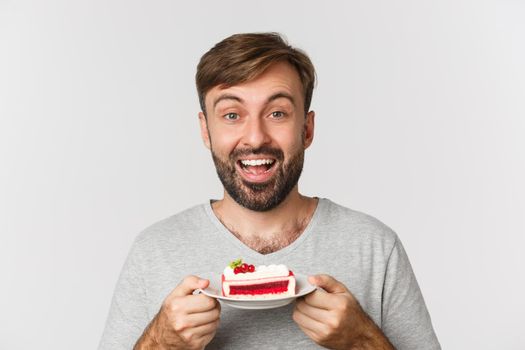 Close-up of handsome smiling man holding cake, standing over white background excited to eat dessert.