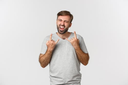 Portrait of carefree bearded man having fun, showing rock-n-roll gesture and shouting for joy, standing over white background.