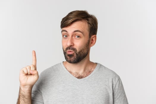 Close-up of thoughtful bearded man in gray t-shirt, having idea, raising finger and looking intrigued, standing over white background.