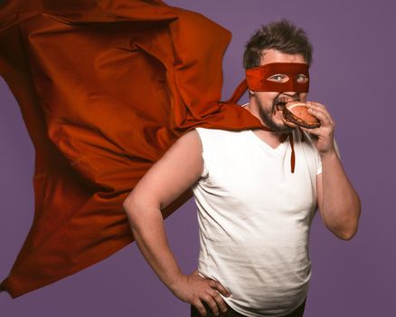 Hungry superhero man eats big hamburger with meat. Man in red flying cloak eats looking at camera on grape purple background. Fast food snack concept.