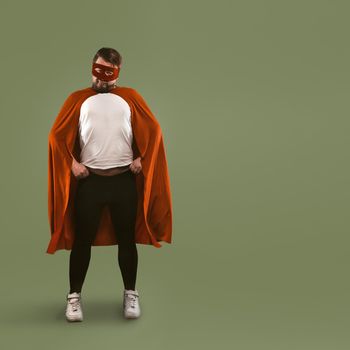 Superhero man stands akimbo at full height. Young Caucasian man in a red superhero costume looks at camera. Isolated on green background. Template with place for text.