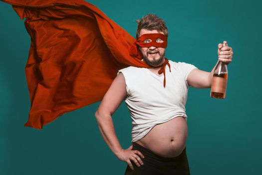 Drunk Superhero Or Antihero Man With Bottle Of Alcohol, Man In Superhero Red Mask And Fluttering Cloak Holding Bottle Of Wine Shows His Big Tummy, Isolated On Biscay Green Background.