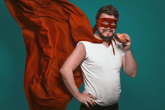 Hungry super hero man eating a hamburger, super hero or antihero in a red suit with a flying cloak eats fast food looking at the camera. Cut out on a green background.