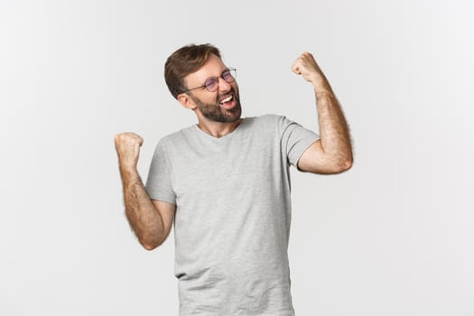 Image of excited man in gray t-shirt and glasses, winning something, rejoicing and feeling like champion, standing over white background.