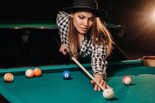 A girl in a hat in a billiard club with a cue in her hands hits a ball.Playing pool.