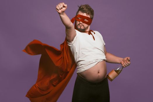 Drunk super antihero holding wine bottle showing his tummy. Man in a red mask and a superhero cloak with bottle of wine in one hand and raising his fist forward. Isolated on grape purple background.