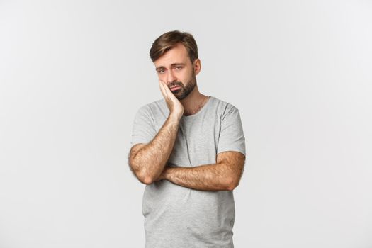 Portrait of sad and bored bearded man in gray t-shirt, leaning on palm and looking reluctant, standing over white background.