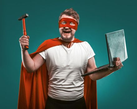 Smiling Super hero man holds hammer and laptop ready to repair the computer. Man In Red Super Hero Uniform posing on biscay green background. Computer repair concept.