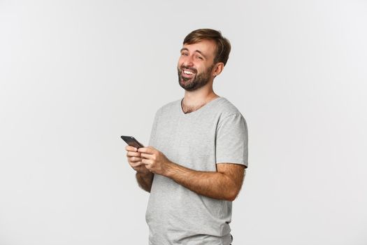 Portrait of caucasian bearded guy in gray t-shirt, laughing at something funny online, holding smartphone, standing over white background.