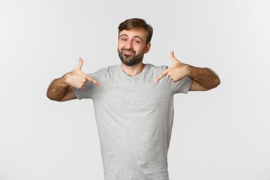 Portrait of handsome bearded man in gray t-shirt pointing at logo and smiling, recommending brand, standing over white background.