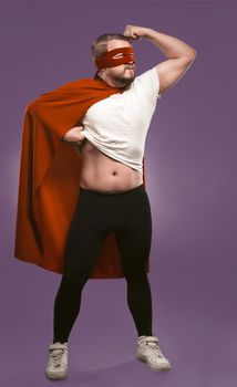 Strong super hero man showing muscles. Brave man in red super hero cape and mask standing full height on grape purple background. Strength concept.
