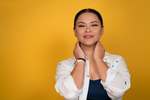 Skin care concept. Pretty woman smiles touching her neck with hands. Copy space at left side. Happy mid aged Asian woman posing in studio on yellow background.