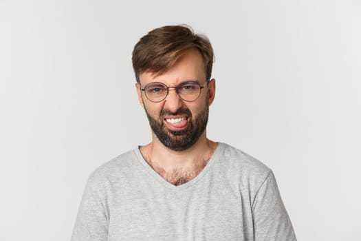 Close-up of skeptical bearded man in glasses and gray t-shirt, grimacing disgusted and showing tongue, standing over white background.