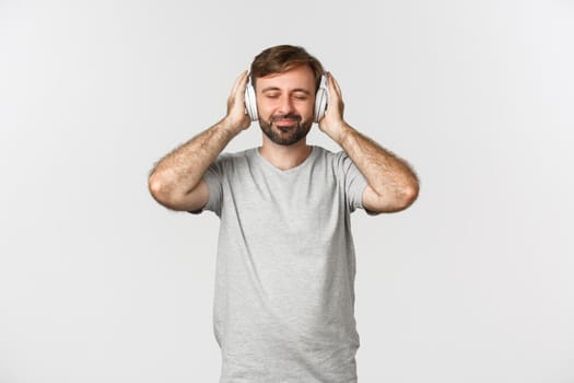 Image of handsome man in casual outfit, listening music in wireless headphones, smiling pleased, standing over white background.