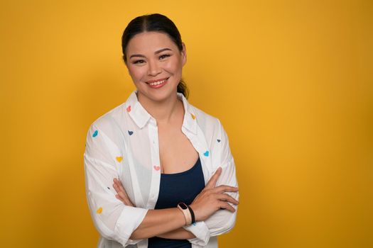 Confident Asian woman smiles standing crossed hands on yellow background. Copy space.