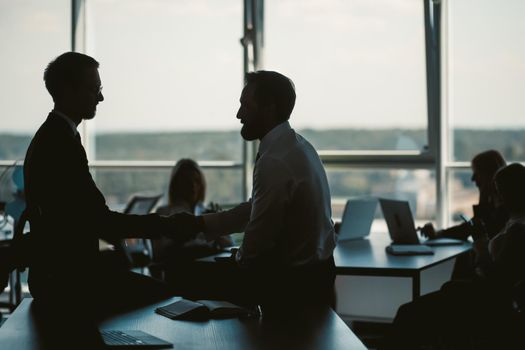 Dark silhouettes of office workers against of window on blurred background. Selective focus on two businessmen communicating while sitting on table in foreground. Toned image.