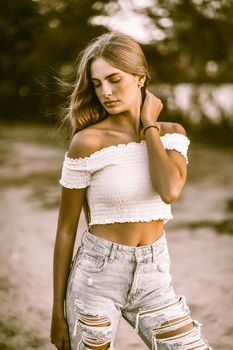 Fashion teenage girl wearing jeans and white top posing in evening on nature background. Vogue concept. Toned image.