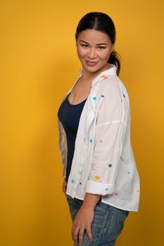 Pretty mature Asian woman wearing jeans posing in studio. Smiling female brunette isolated on yellow background.