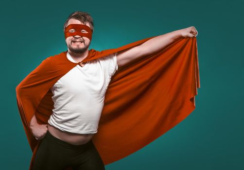 Funny Super Hero Man Ready To Fly Save World. Smiling Man In Mask And Red Superhero Costume Holding His Cloak In His Hand Posing On Biscay Green Background.