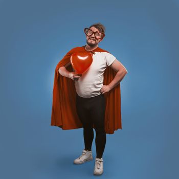 Romantic super hero holds big heart. Happy man in love wearing superhero costume and heart-shaped glasses isolated on blue background. Valentine's day concept. Copy spase on both sides.