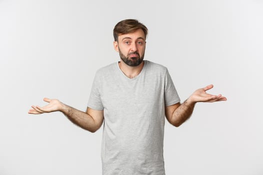 Image of clueless bearded man in casual t-shirt, shrugging and looking puzzled, standing over white background.