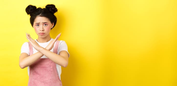 Sad girl begging to stop, frowning uspet and showing cross sign, say no, standing gloomy on yellow background.