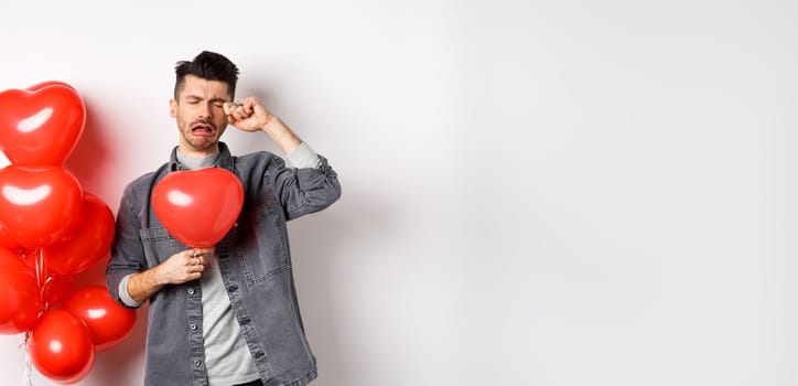 Valentines day and love concept. Sad crying man holding red heart balloon and whiping tears, standing single and miserable, being heartbroken, white background.