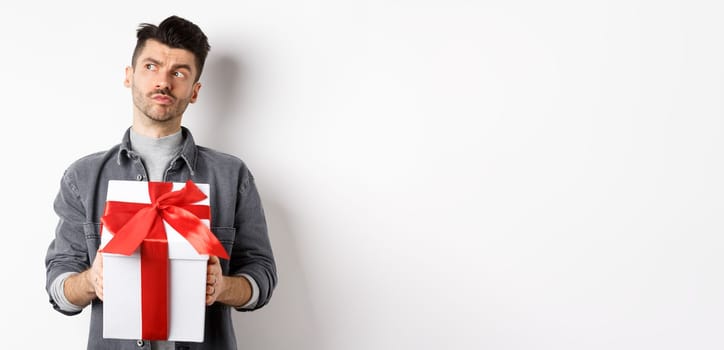 Pensive boyfriend looking aside and holding gift box, waiting for lover, making surprise present on valentines day, planning date with girlfriend, white background.