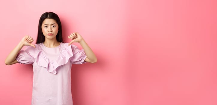 Skeptical and disappointed asian woman showing thumbs down with reluctant smirk, dislike product, show negative emotion, standing on pink background.