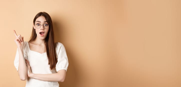 Creative young woman in glasses pitching idea, raising finger in eureka sign and gasping, have a plan, standing on beige background.