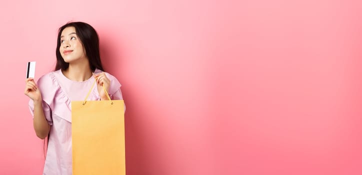 Shopping. Dreamy asian girl thinking of buying something, holding shop bag and plastic credit card, standing against pink background.