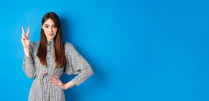 Young beautiful girl in dress with natural long hair, showing number two with fingers and smiling, standing against blue background.