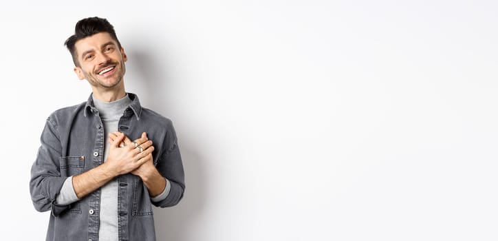 Romantic guy hold hands on heart and smiling, thanking you, looking pleased and grateful, standing on white background.