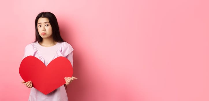 Valentines concept. Single teenage asian girl wants to fall in love, looking sad and lonely at camera, sulking distressed on lovers day, holding big red heart cutout, pink background.