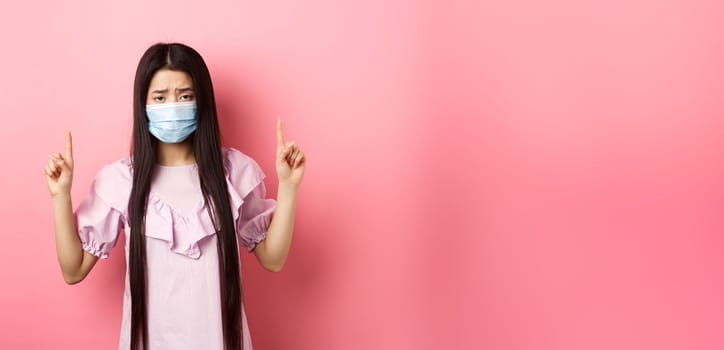 Healthy people and covid-19 pandemic concept. Sad asian girl feeling sick, wearing medical mask on self-quarantine, standing against pink background.