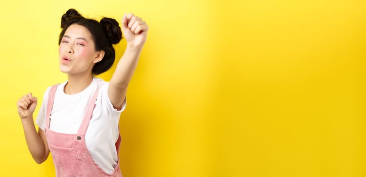 Excited asian girl looking motivated, raising hand up and chanting, cheering with joy, standing happy on yellow background.