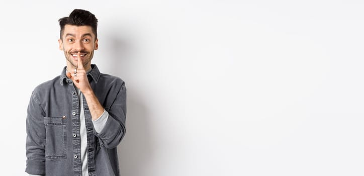 Happy guy shushing at camera, smiling with finger on lips, asking to keep quiet, telling big secret or making surprise, standing on white background.