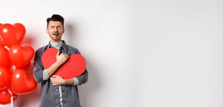 Crying man standing single and lonely on Valentines day, hugging heart cutout and sobbing miserable, being heartbroken and rejected by lover, white background.