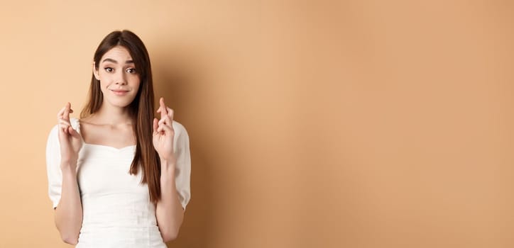 Hopeful tender girl in white blouse making wish, holding fingers crossed and smiling, praying for good luck, waiting for results, beige background.
