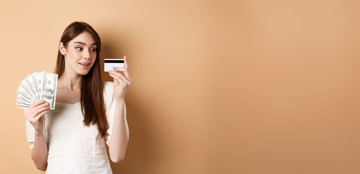 Dreamy girl looking at plastic credit card and thinking of shopping, holding dollar bills, standing on beige background.