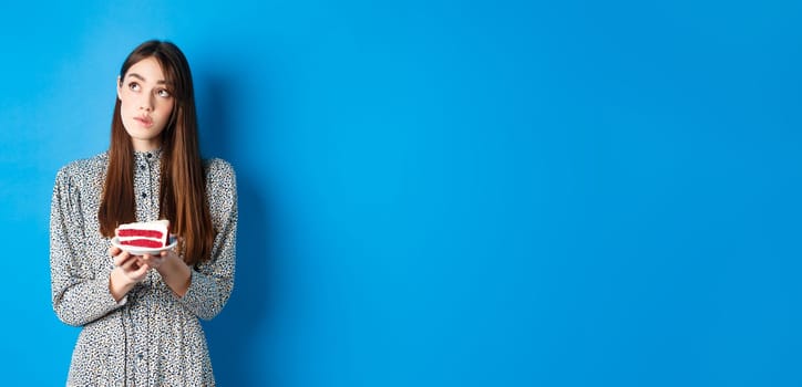 Birthday girl making wish, looking aside at logo and holding b-day cake, standing on blue background.