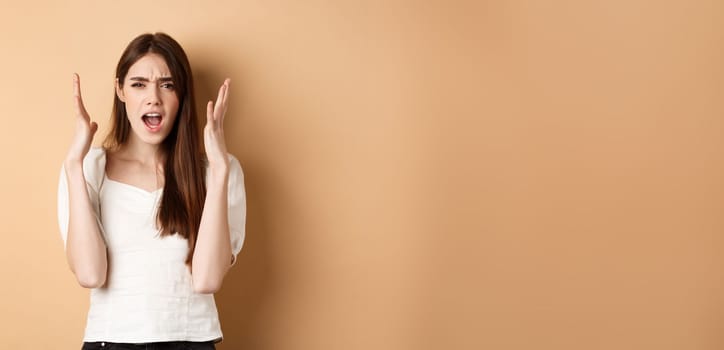 Frustarted woman shouting and shaking hands angry, looking mad, standing distressed on beige background.