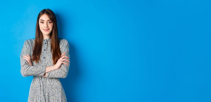 Elegant caucasian girl in dress cross arms on chest, standing on blue background.