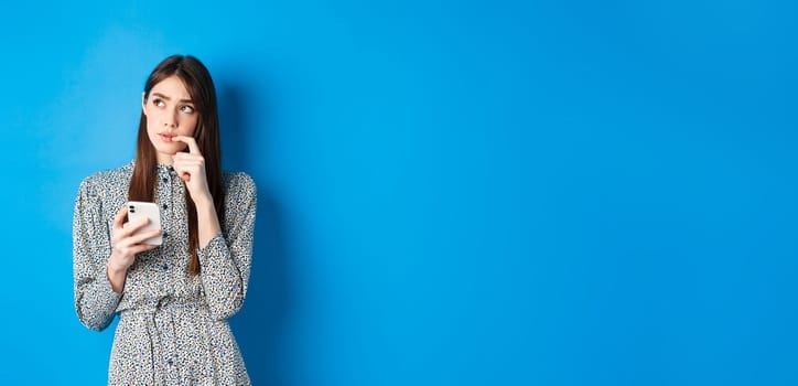 Pensive cute girl thinking how to answer on message, looking aside thoughtful and holding smartphone, standing in dress on blue background.