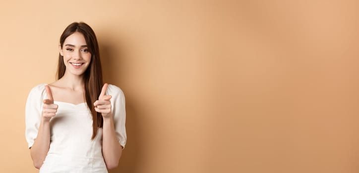 We need you. Smiling young woman pointing fingers at camera to beckon or invite people, standing on beige background.