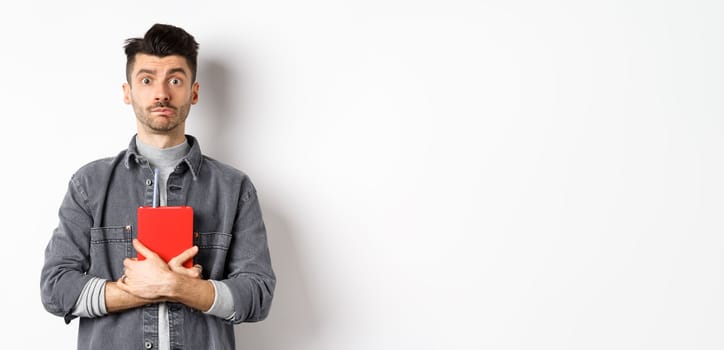 Cute and shy guy hugging his diary, holding red journal and look modest at camera, standing against white background.